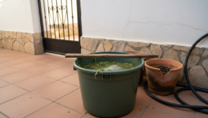 Cubo de plástico con purín casero en fermentación junto a macetas de barro en un patio español a la sombra.